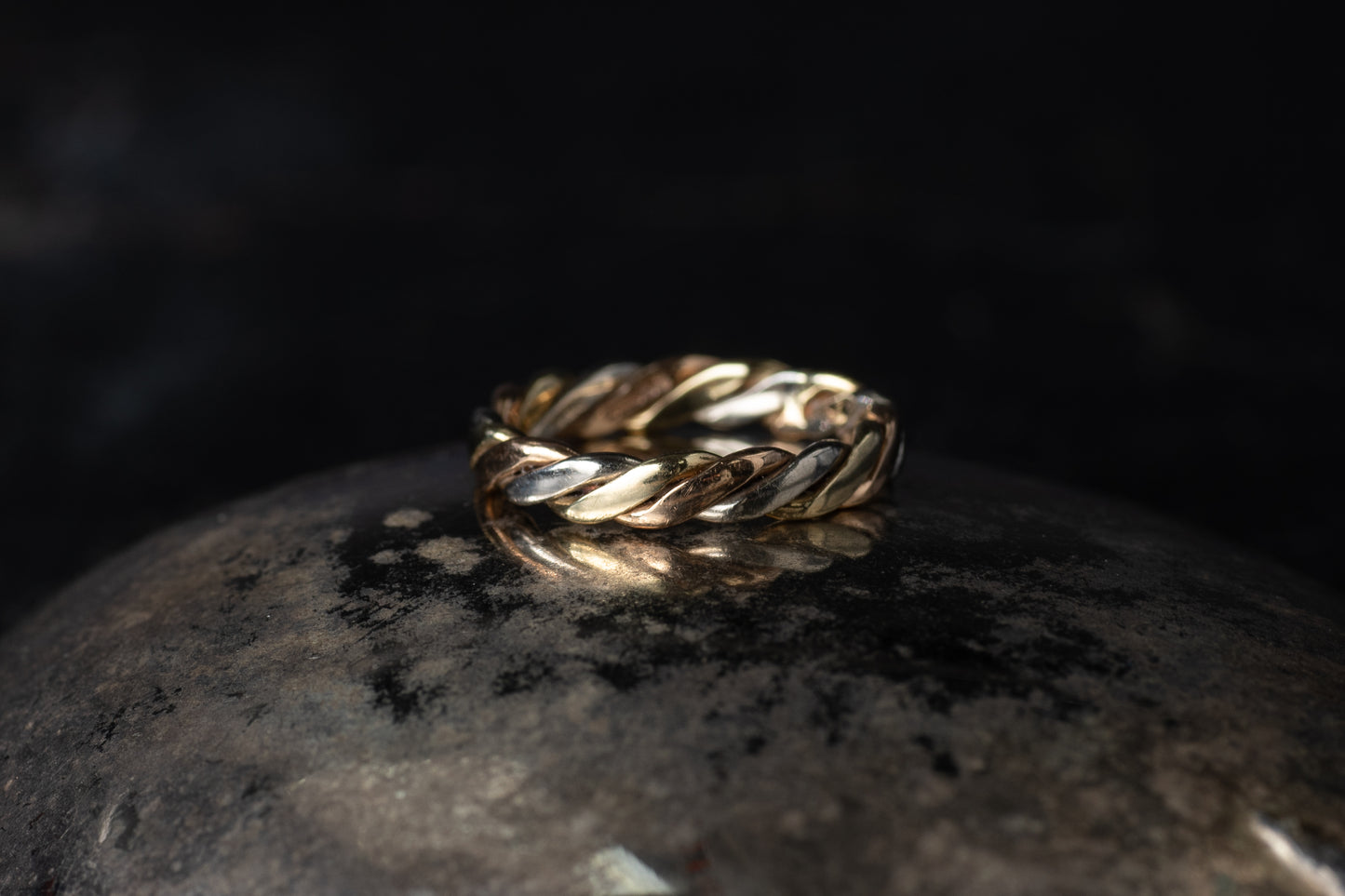 9ct gold ring featuring intertwined bands of yellow, white, and rose gold in a twist design on a dark reflective surface.