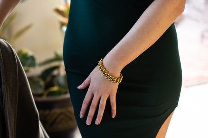 A close-up lifestyle shot of a person wearing a chunky gold chain bracelet on their hand, dressed in a dark green bodycon dress.