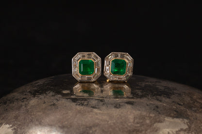 Close up of octagonal 1.25ct emerald and baguette diamond cluster stud earrings set in 18ct yellow gold on metallic surface.