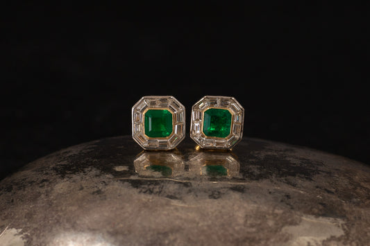 Close up of octagonal 1.25ct emerald and baguette diamond cluster stud earrings set in 18ct yellow gold on metallic surface.
