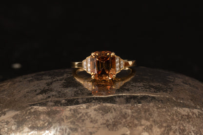 18ct yellow gold ring featuring emerald cut peach tourmaline and stepped diamond shoulders on a dark reflective background.