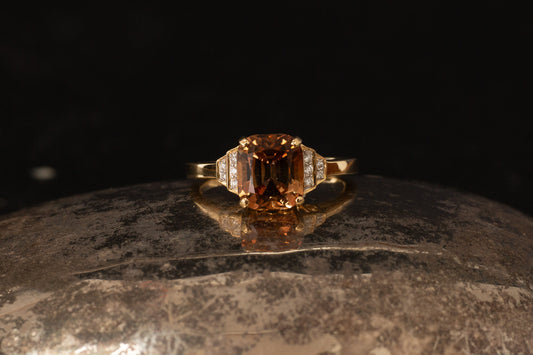 18ct yellow gold ring featuring emerald cut peach tourmaline and stepped diamond shoulders on a dark reflective background.