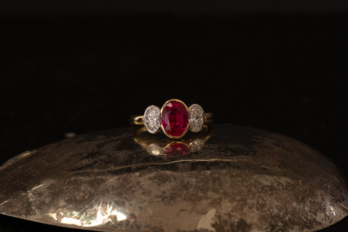 18ct yellow gold three stone ring with oval ruby and side diamonds in rub over setting on dark background.