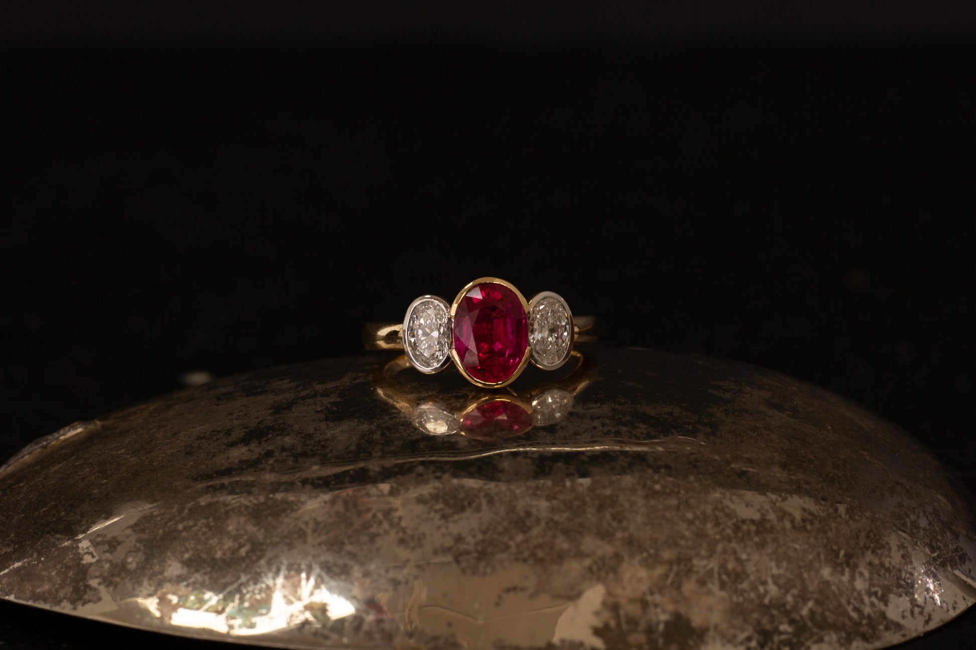 18ct yellow gold three stone ring with oval ruby and side diamonds in rub over setting on dark background.