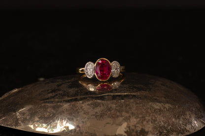 18ct yellow gold three stone ring with oval ruby and side diamonds in rub over setting on dark background.