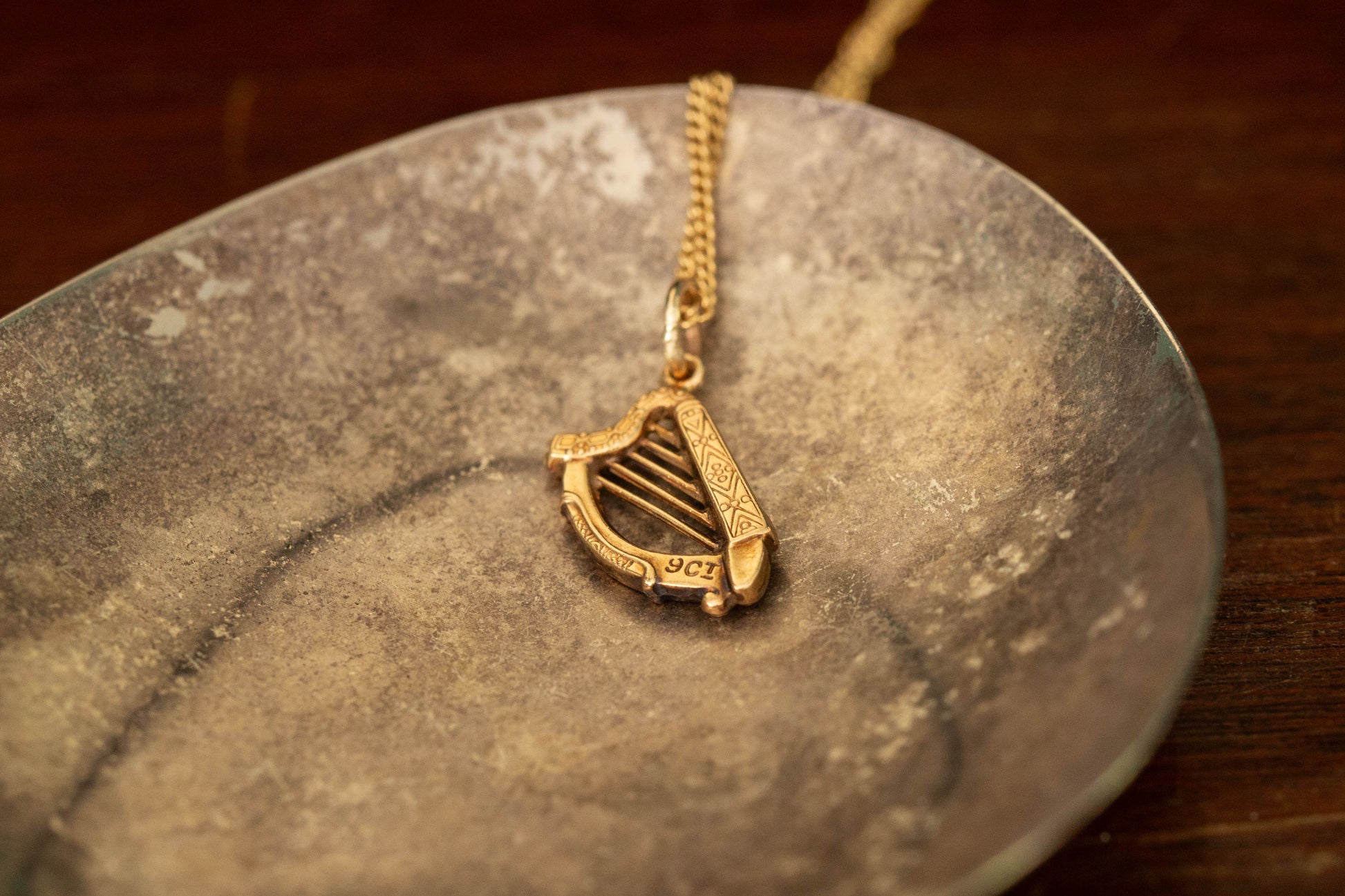 9ct yellow gold harp pendant charm with engraved details lying on a dark metallic dish.