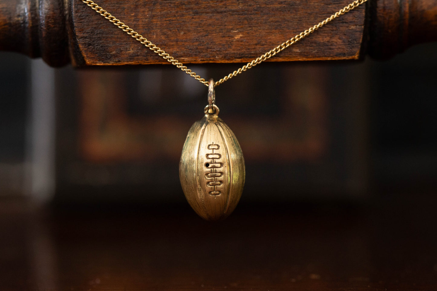 Close up of 9ct yellow gold rugby ball pendant charm with engraved stitching details hanging against a dark background.