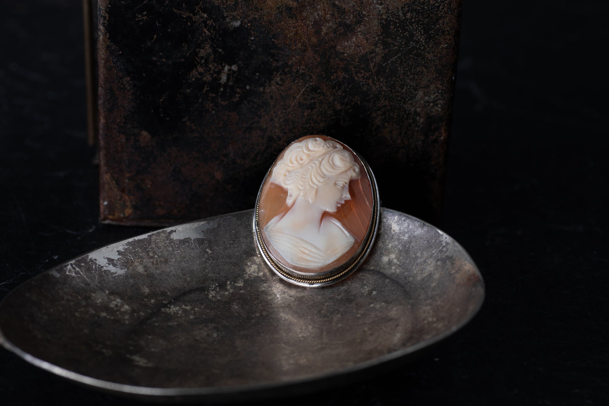 Oval vintage mother of pearl cameo brooch featuring a female profile rub-over set in silver on a dark metallic dish.