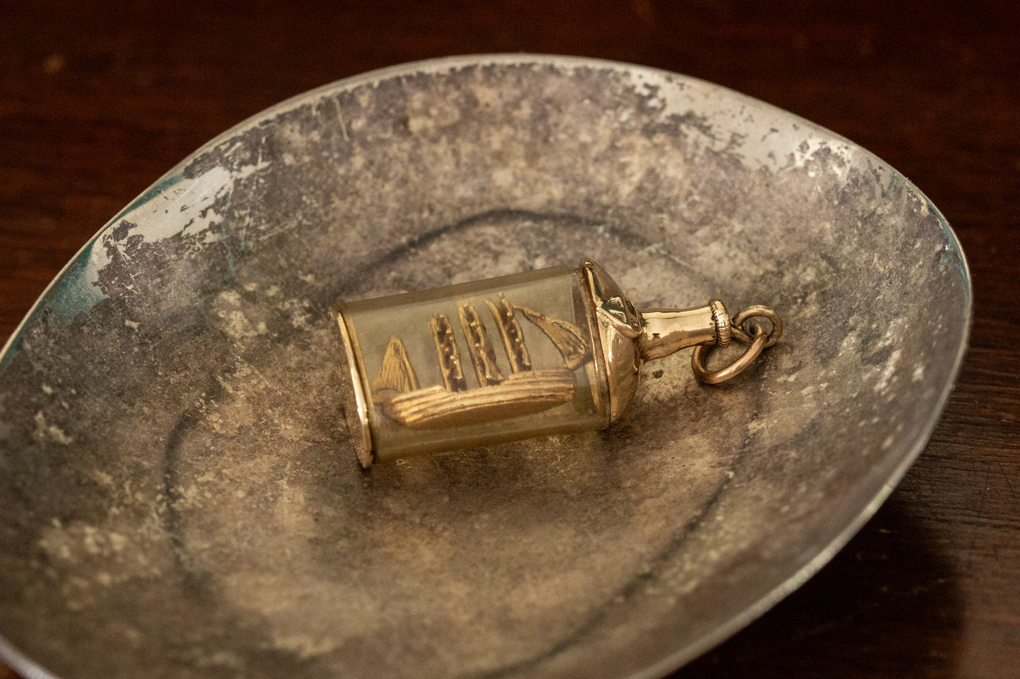 9ct yellow gold and glass ship in a bottle pendant charm lying on a dark metallic dish.