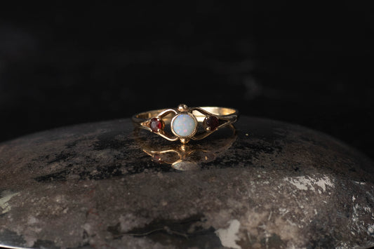 9ct yellow gold vintage ring featuring central round opal and garnet side stones with split shoulders on dark surface.