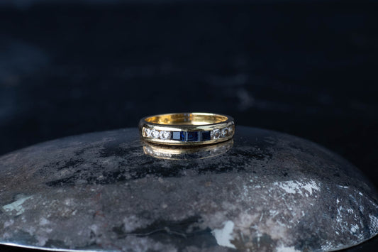 18ct yellow gold half eternity band featuring channel set baguette sapphires and diamonds on a dark reflective surface.
