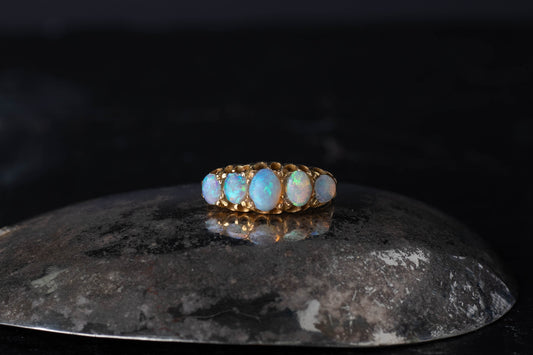 18ct yellow gold antique ring featuring five graduated claw set opals on a dark reflective surface.
