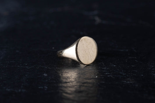9ct yellow gold signet ring featuring a plain oval face resting on a dark textured surface.