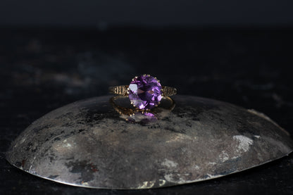 9ct yellow gold vintage ring featuring a round solitaire amethyst claw set with textured shoulders on a dark reflective surface.