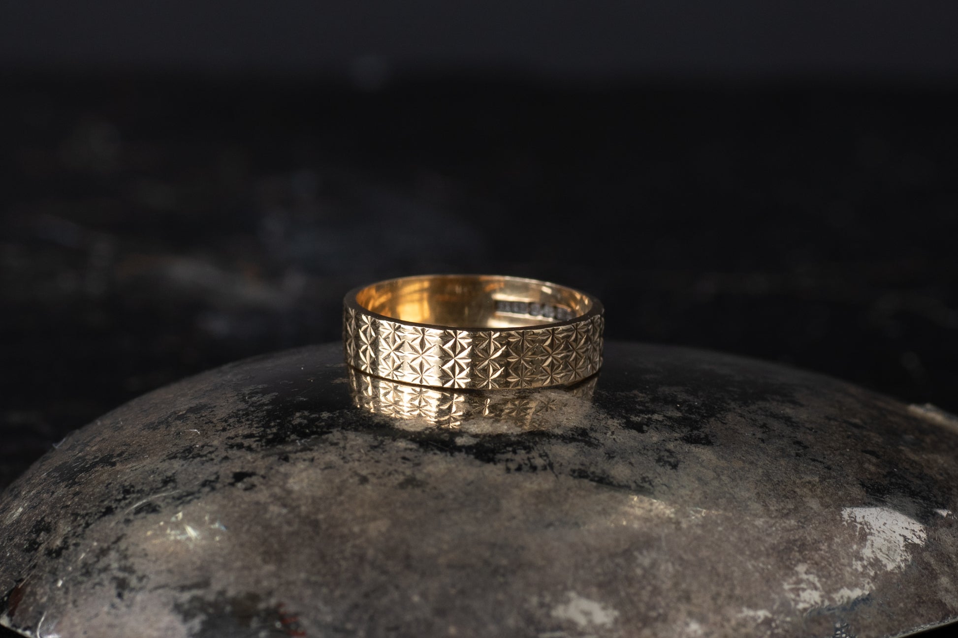 Gold wedding band ring featuring an intricate engraved geometric pattern resting on a dark reflective surface.