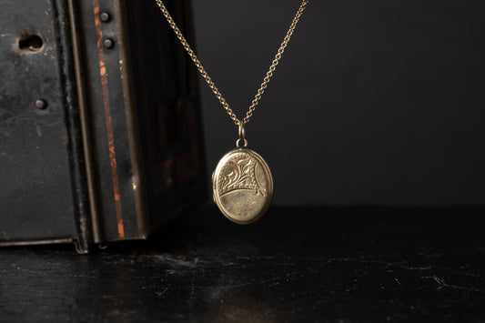 Gold plated oval locket with engraved corner flourish design hanging against a dark background.
