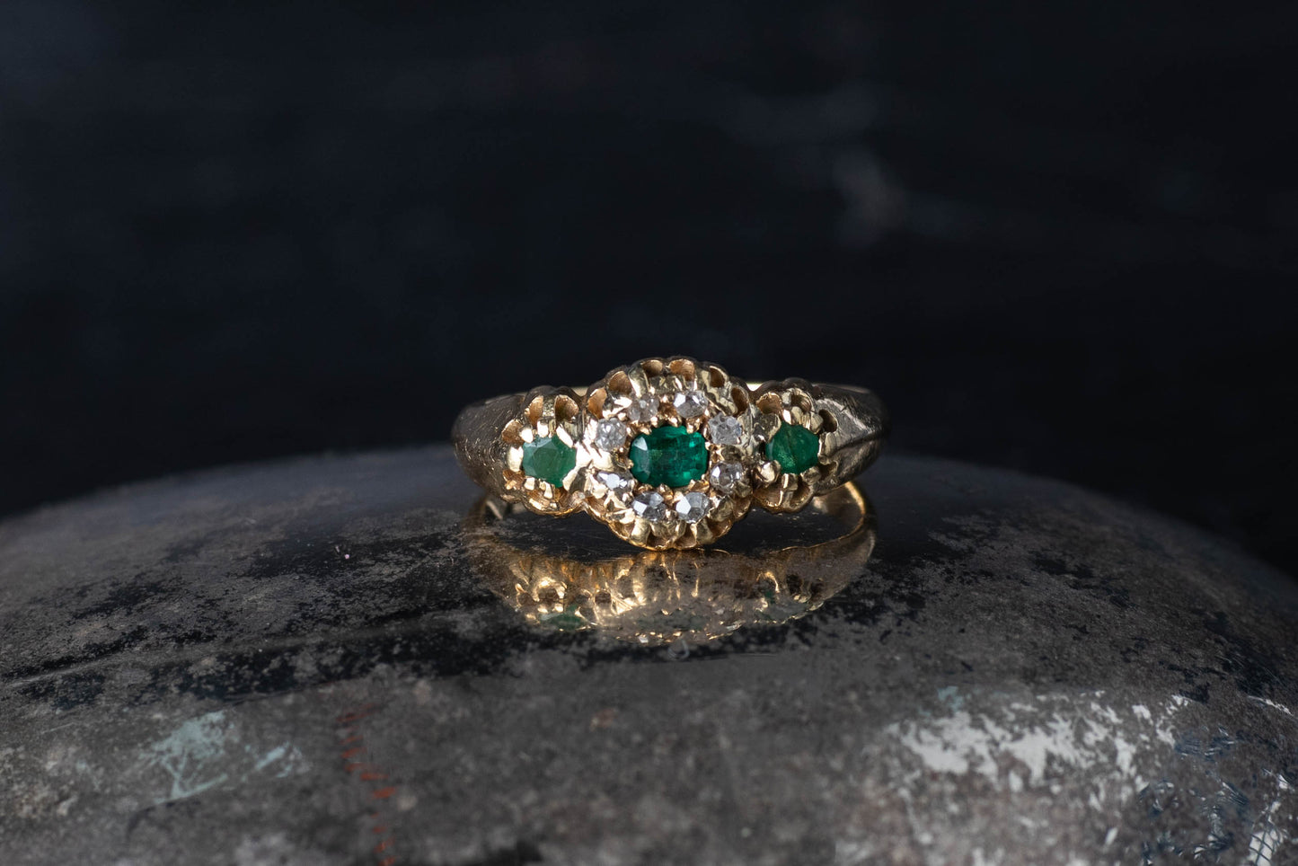 Vintage 18ct gold ring featuring a central emerald and diamond cluster with emerald shoulders on a dark reflective surface.