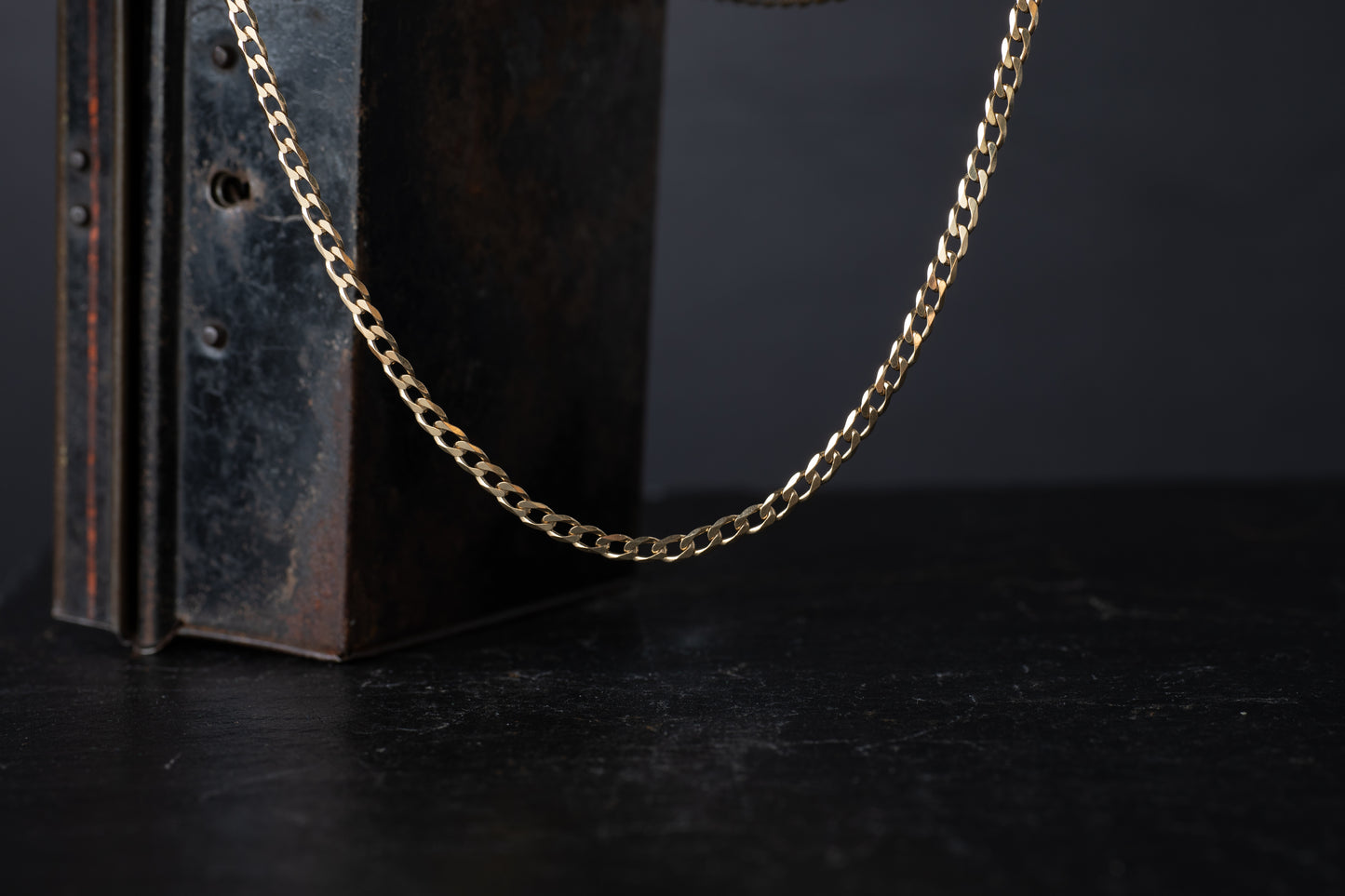 9ct yellow gold flat bevelled curb link chain necklace hanging against a dark vintage box background.