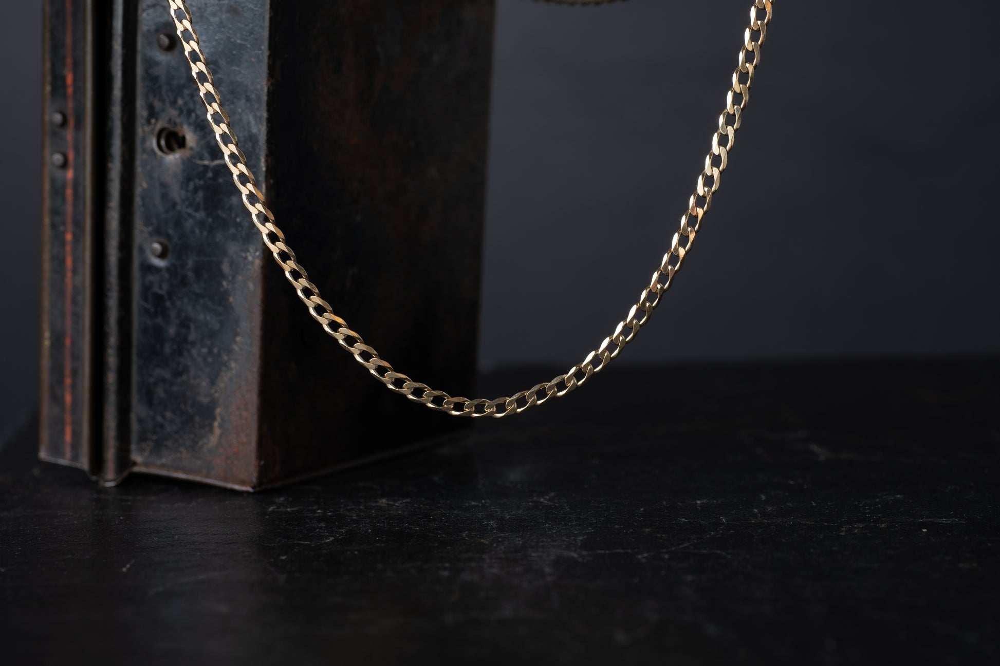 9ct yellow gold flat bevelled curb link chain necklace hanging against a dark vintage box background.