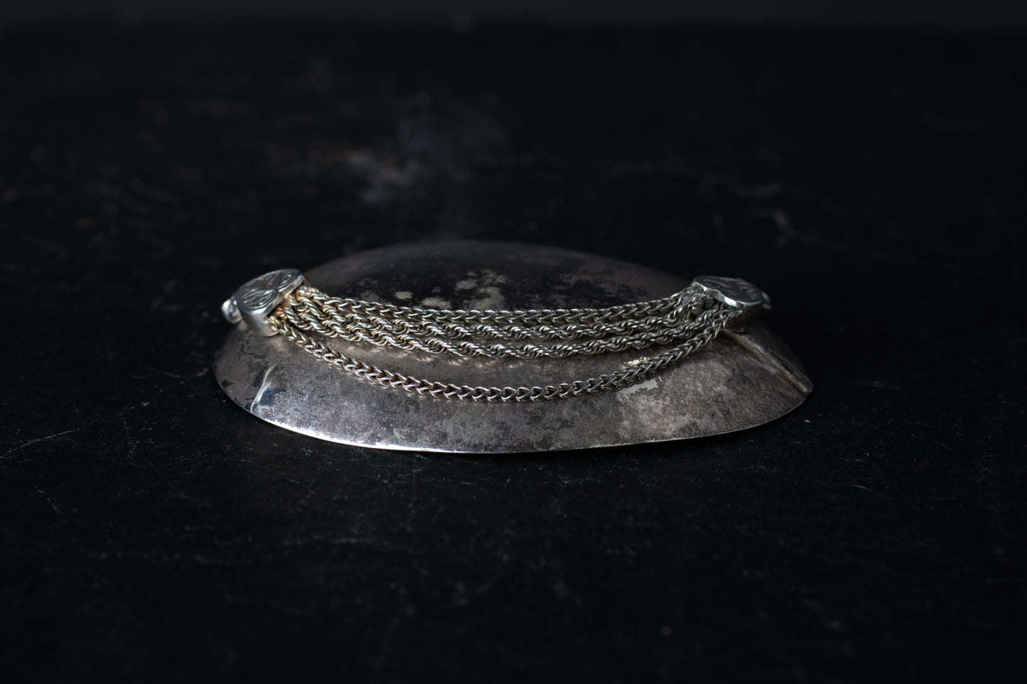 Antique silver bracelet featuring multiple mixed link chains connected to heart-shaped terminals on a dark reflective surface.