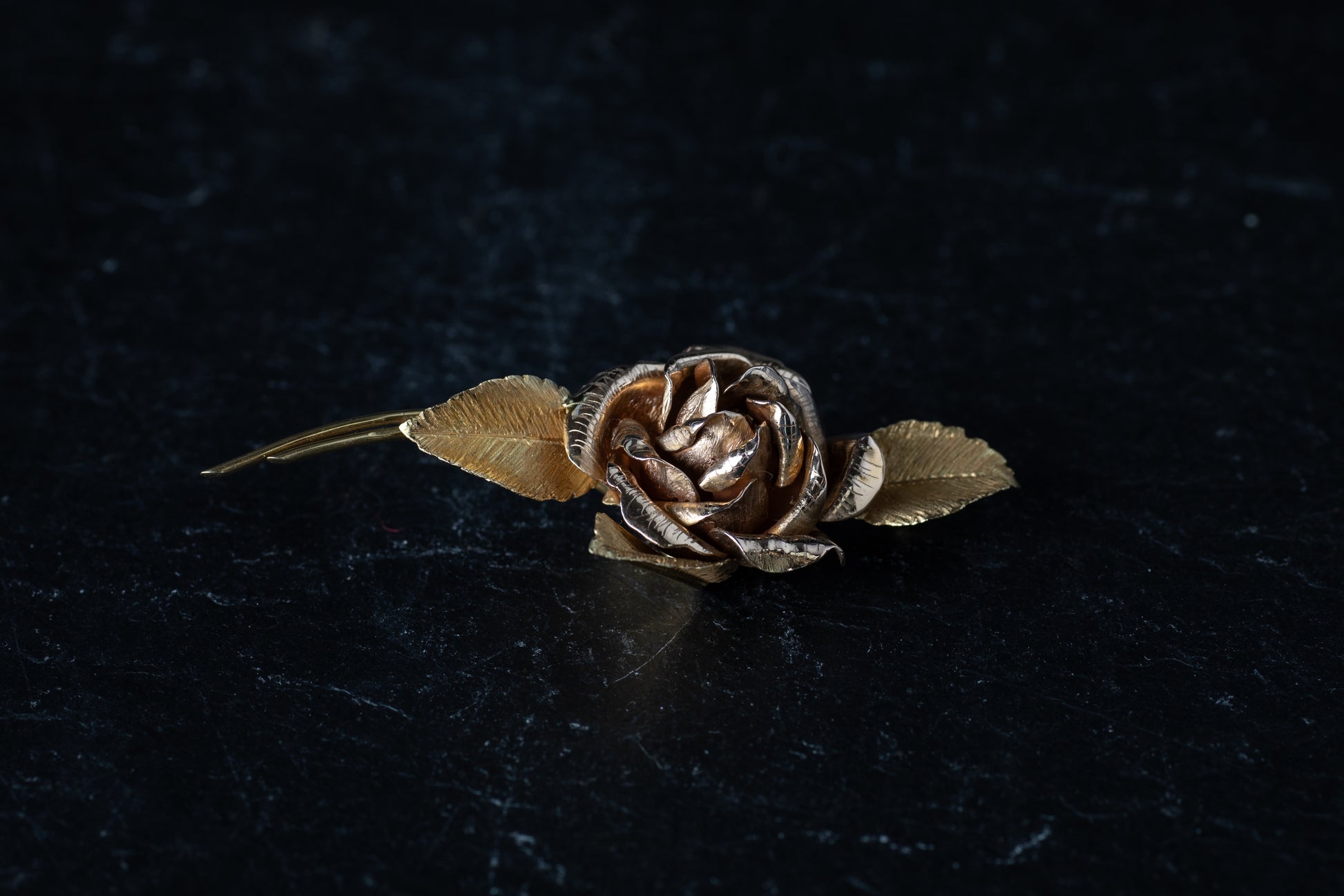18ct yellow gold bar brooch designed as a blooming rose with textured leaves resting on a dark reflective surface.