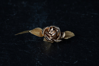18ct yellow gold bar brooch designed as a blooming rose with textured leaves resting on a dark reflective surface.