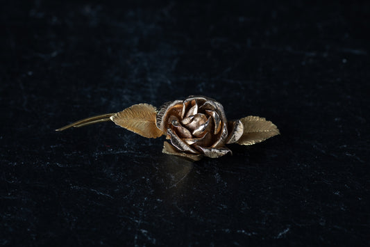 18ct yellow gold bar brooch designed as a blooming rose with textured leaves resting on a dark reflective surface.