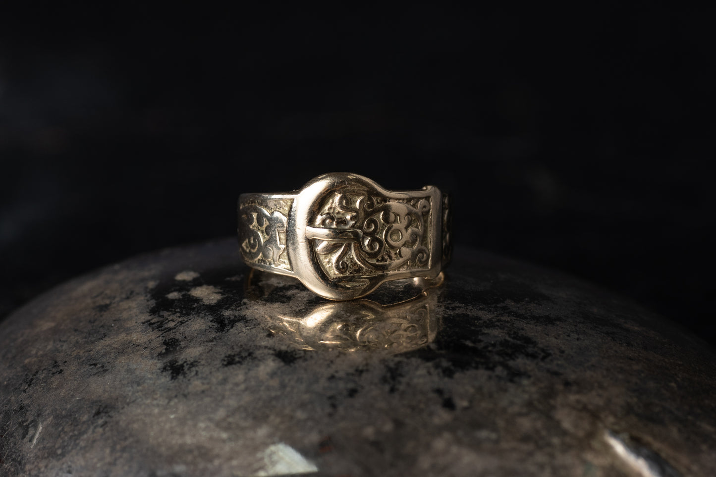 Close up of vintage 9ct yellow gold ring with belt buckle design and engraved scrollwork on dark background.