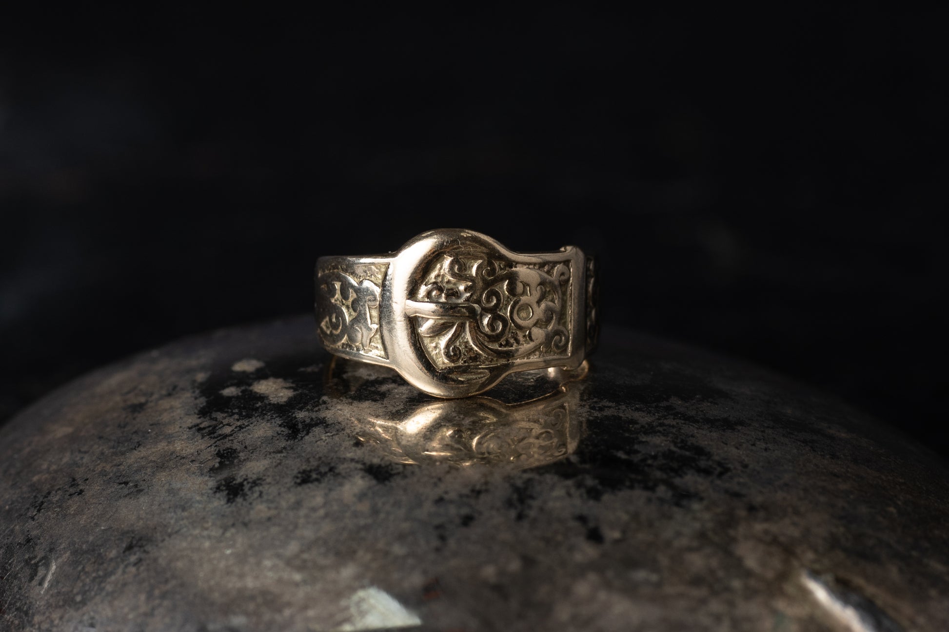 Close up of vintage 9ct yellow gold ring with belt buckle design and engraved scrollwork on dark background.