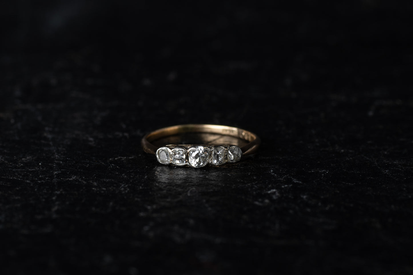 Front view of antique five stone diamond ring with platinum rub over setting on a yellow gold band against a dark textured background.