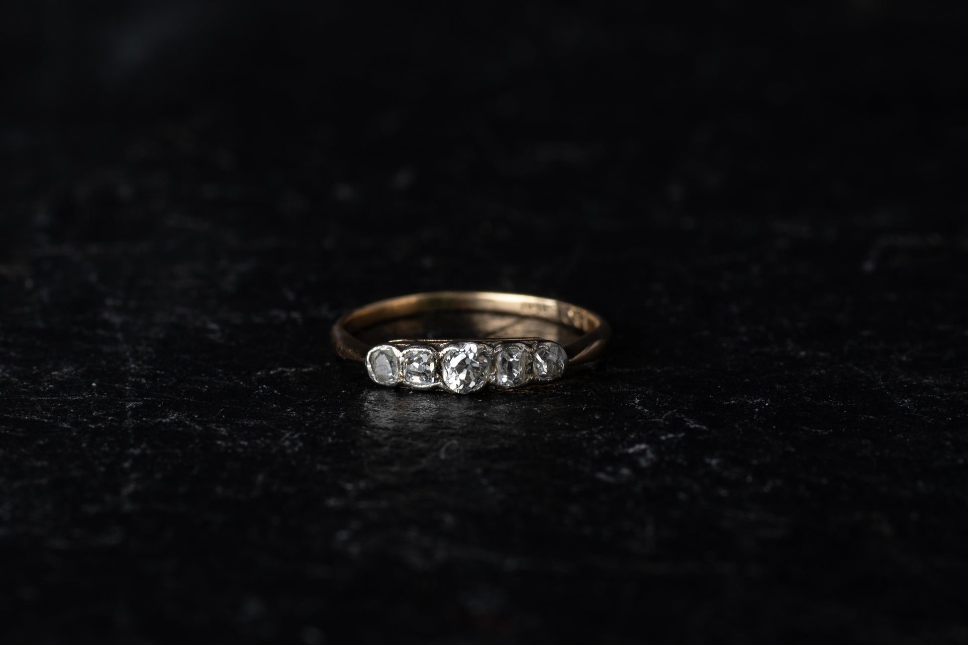 Front view of antique five stone diamond ring with platinum rub over setting on a yellow gold band against a dark textured background.