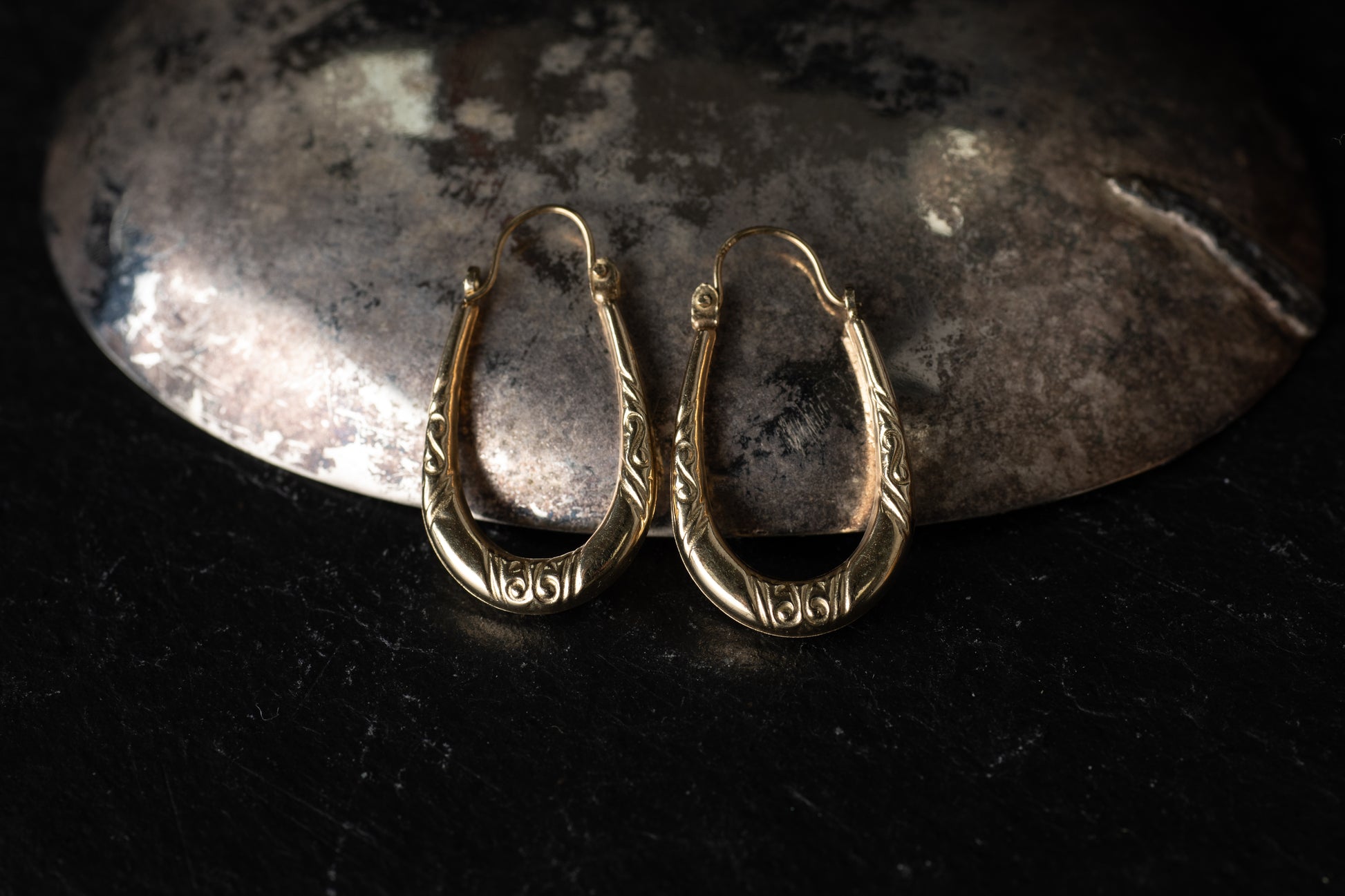 Pair of 9ct yellow gold creole hoop earrings with engraved scroll details resting on dark stone background.