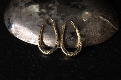Pair of 9ct yellow gold creole hoop earrings with engraved scroll details resting on dark stone background.