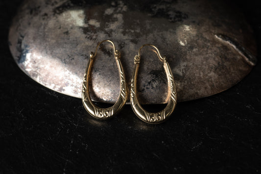 Pair of 9ct yellow gold creole hoop earrings with engraved scroll details resting on dark stone background.