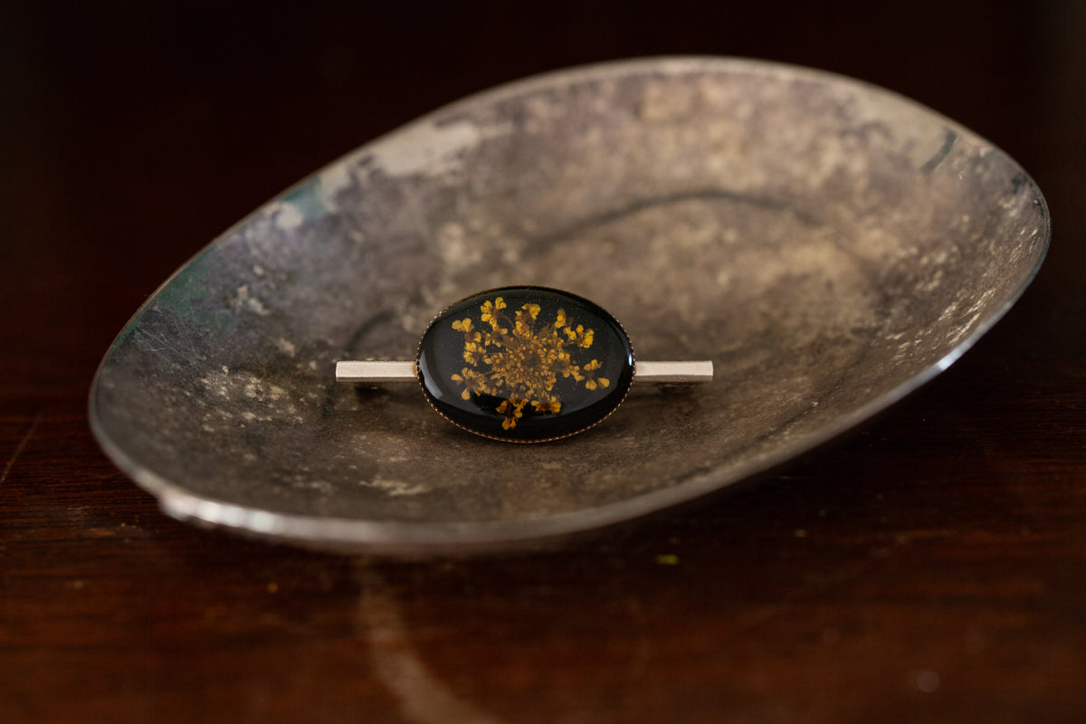 Oval pressed flower brooch with yellow florals on black background rub over set on silver bar pin.