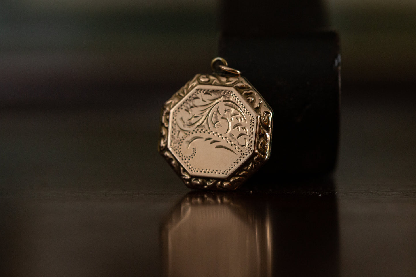 Front view of 9ct rose gold hexagonal locket with vintage floral engraving standing on a dark reflective surface.