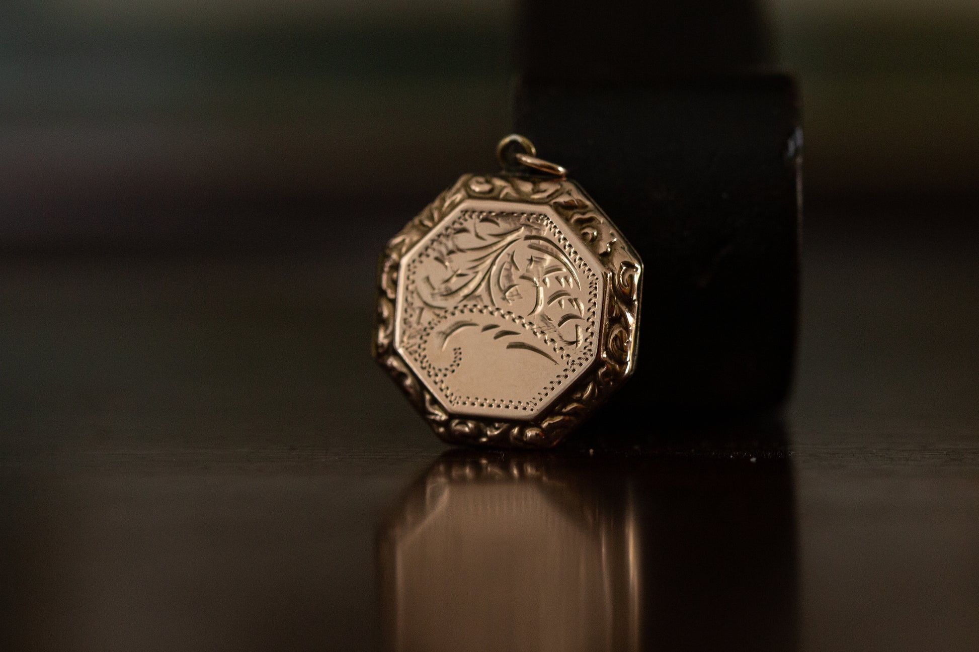 Front view of 9ct rose gold hexagonal locket with vintage floral engraving standing on a dark reflective surface.