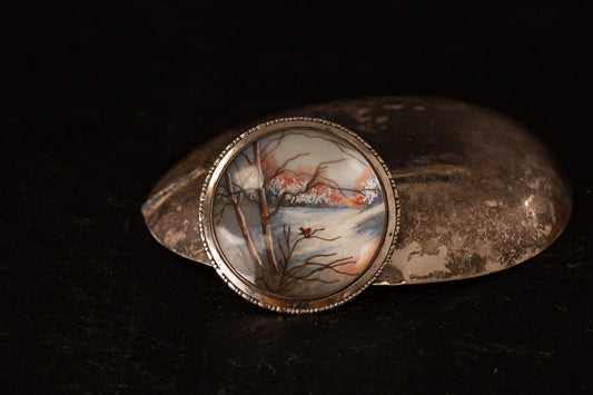 Round vintage silver enamel brooch featuring hand-painted winter landscape with trees and snow on dark background.