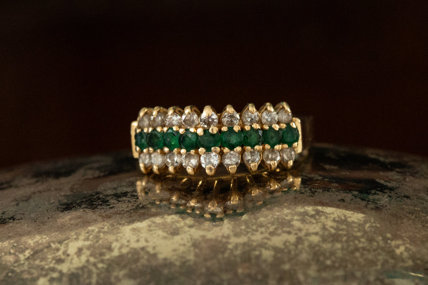Front view of 9ct yellow gold triple row ring with central emeralds and outer diamond rows claw set on dark prop.