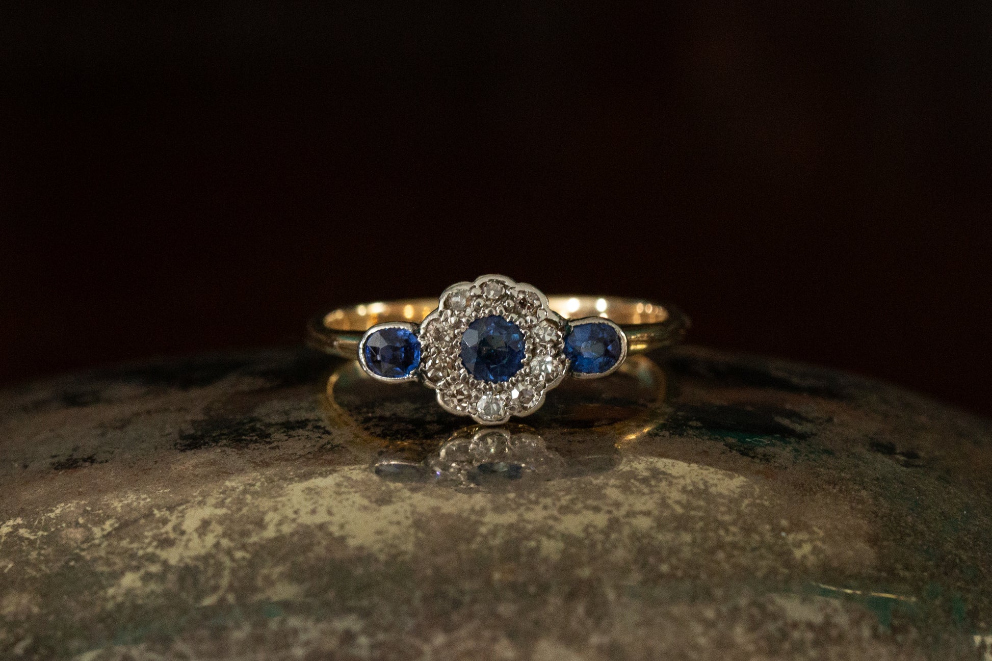 Front view of Victorian sapphire and diamond cluster ring featuring rub over set side stones on a dark reflective background.