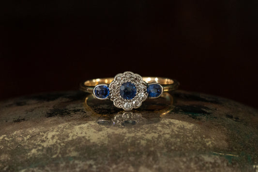 Front view of Victorian sapphire and diamond cluster ring featuring rub over set side stones on a dark reflective background.