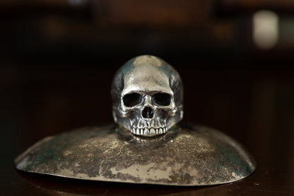Front view of realistic solid silver skull ring featuring detailed teeth and eye sockets on a dark reflective background.