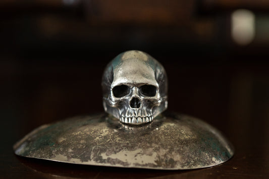 Front view of realistic solid silver skull ring featuring detailed teeth and eye sockets on a dark reflective background.