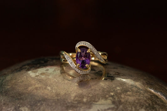 Front view of yellow gold twist ring featuring an oval amethyst and diamond bypass setting on a dark reflective background.