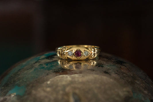 Front view of Victorian 18ct yellow gold ring featuring a central ruby and two diamonds with ornate scroll shoulders on dark background.