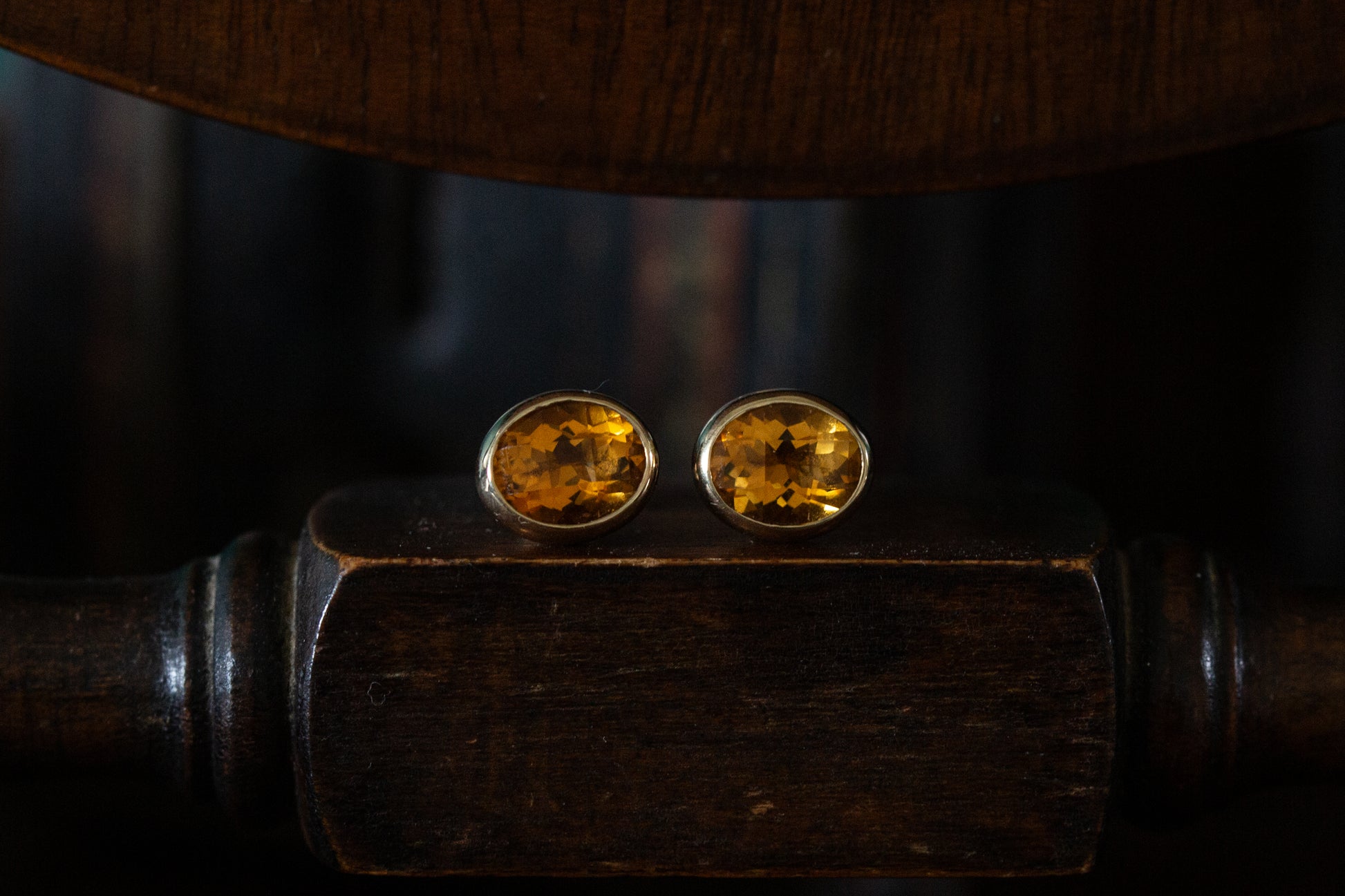 Front view of large faceted oval citrine stud earrings rub over set in 9ct yellow gold resting on a dark wooden surface.