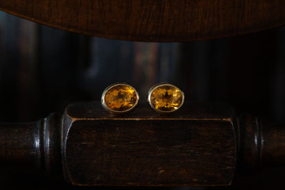 Front view of large faceted oval citrine stud earrings rub over set in 9ct yellow gold resting on a dark wooden surface.