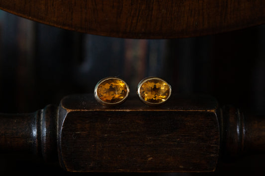 Front view of large faceted oval citrine stud earrings rub over set in 9ct yellow gold resting on a dark wooden surface.
