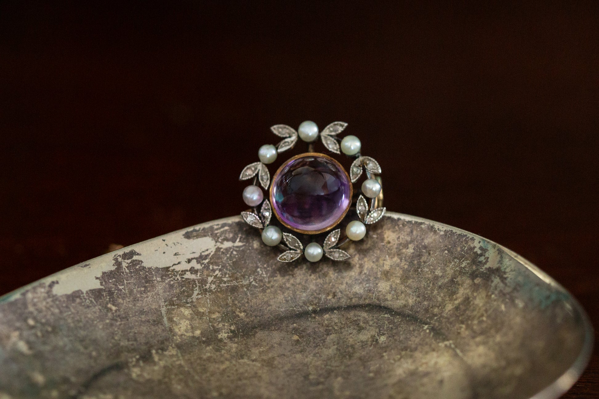 Front view of antique round brooch featuring a central cabochon amethyst surrounded by a wreath of rose cut diamonds and seed pearls in 18ct rose gold.