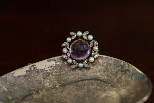 Front view of antique round brooch featuring a central cabochon amethyst surrounded by a wreath of rose cut diamonds and seed pearls in 18ct rose gold.